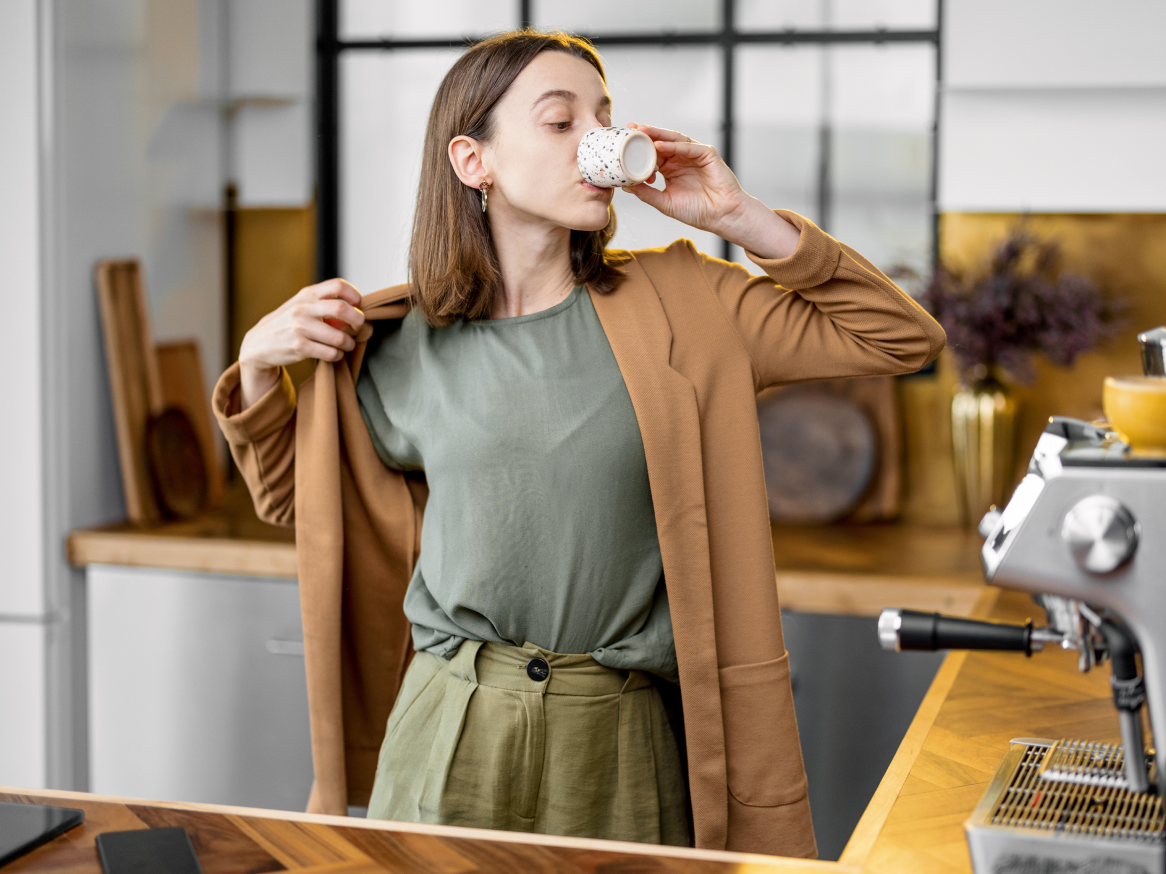 Mulher em uma cozinha moderna, vestindo um blazer marrom e calças verdes, bebendo café.