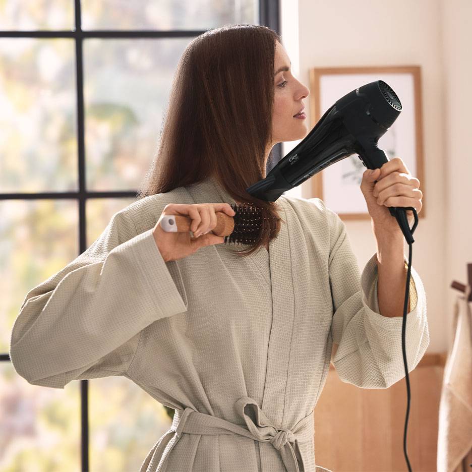 Mulher a secar o cabelo com secador e escova redonda, vestindo um roupão.