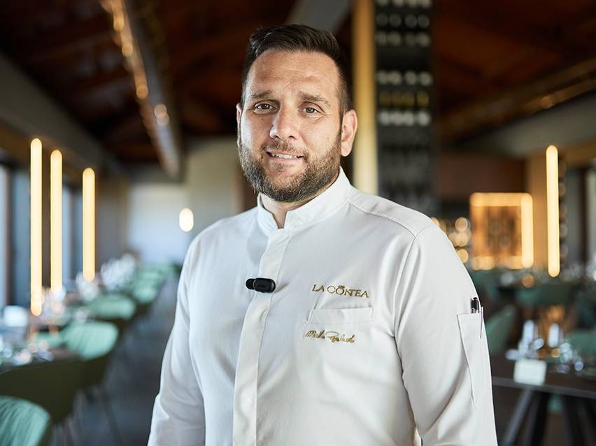 Chef sorridente em uniforme branco com o logótipo 'LA CONTEA' num restaurante elegante.