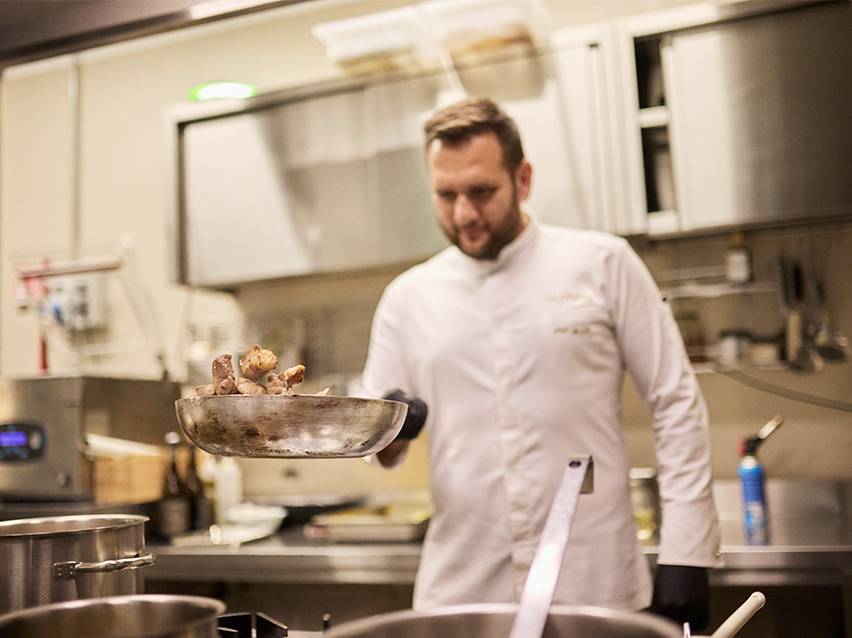 Um chef de cozinha de jaleco branco salteando comida numa frigideira.
