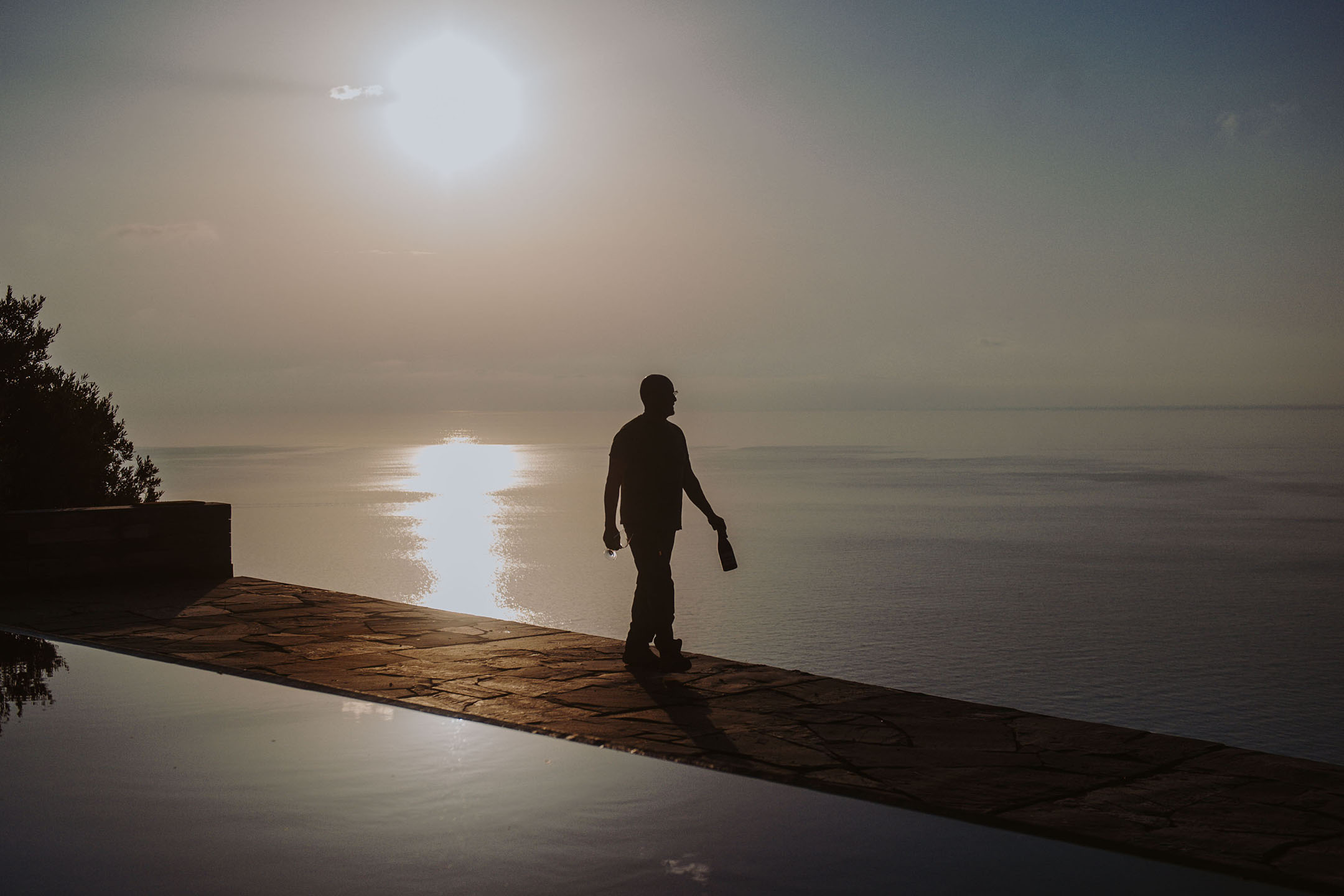 Silhueta de uma pessoa com garrafa e copo junto a piscina infinita ao pôr do sol