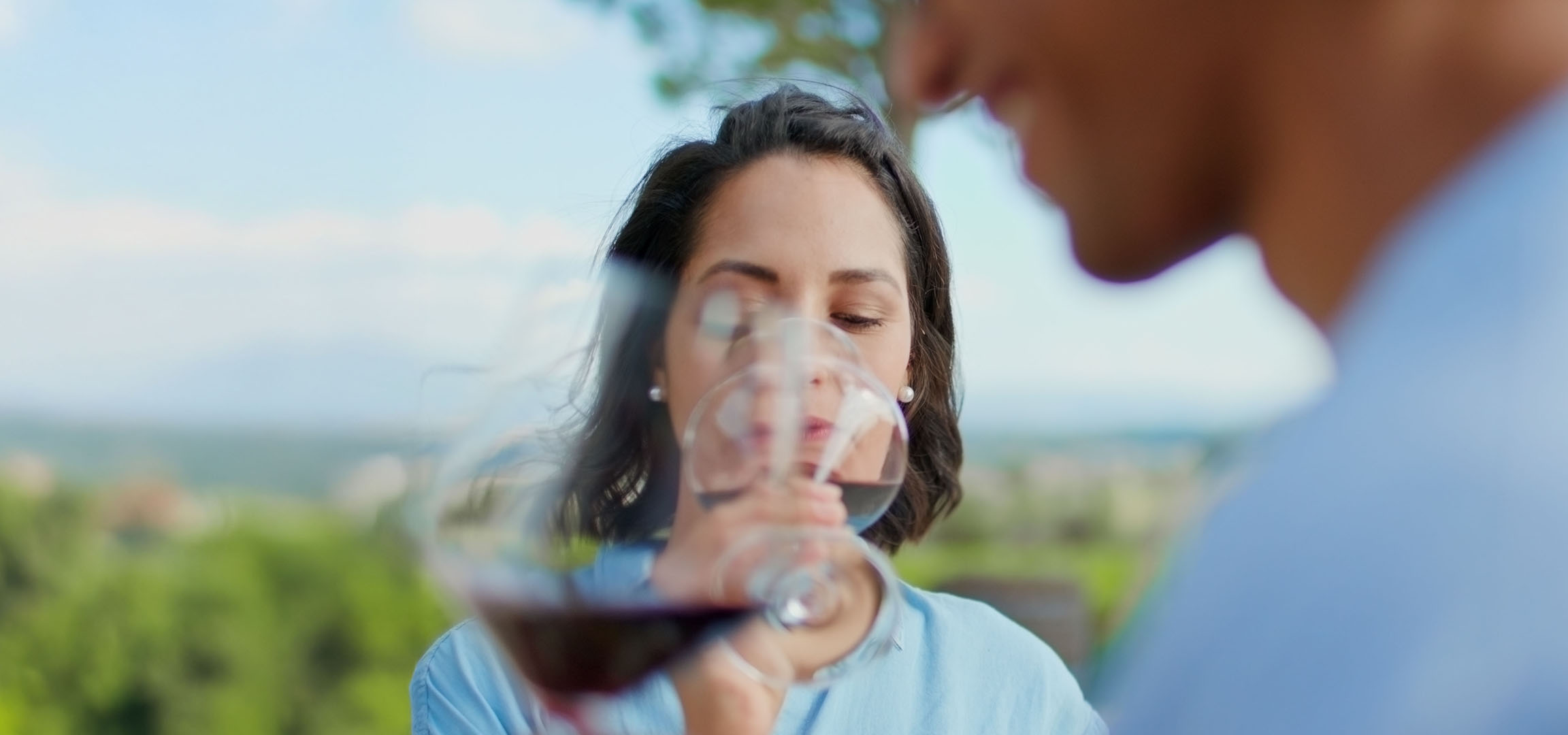 Mulher a desfrutar de vinho tinto ao ar livre, homem em segundo plano.