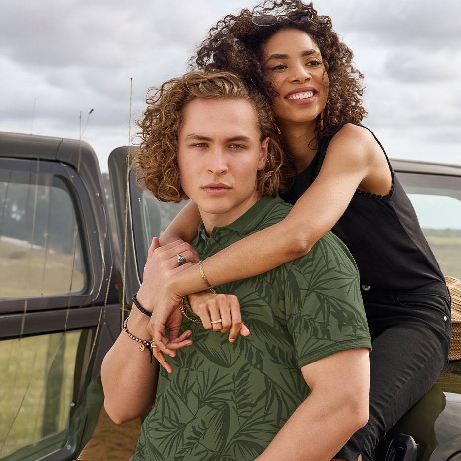 Casal sorridente, ele de polo verde com folhas, ela de top preto, junto a um carro.