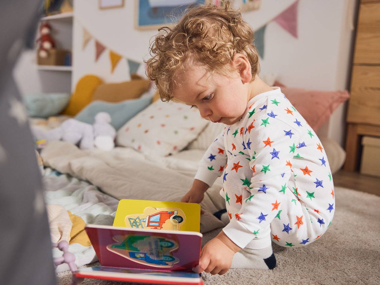 Uma criança pequena de pijama de estrelas, sentada no tapete, lendo um livro.