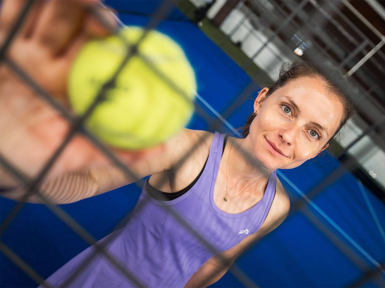 Mulher em roupa desportiva segura bola de padel através da rede, campo azul ao fundo.