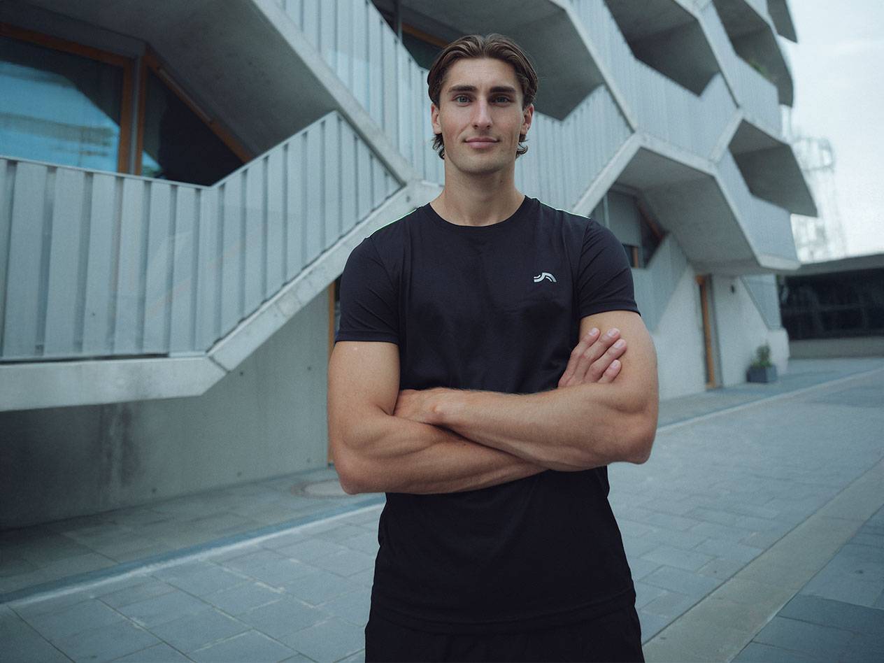 Homem de t-shirt preta com logótipo e calções pretos, de pé ao ar livre com os braços cruzados.