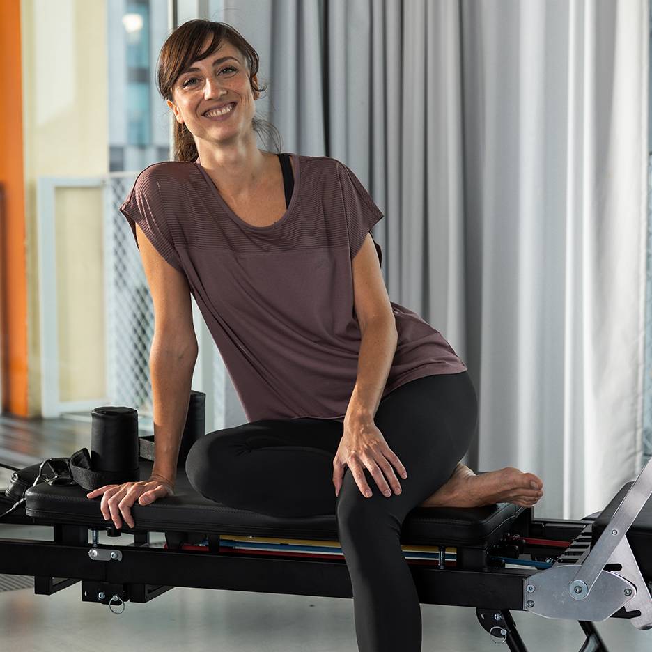 Mulher em roupa desportiva a sorrir, sentada numa máquina de Pilates.