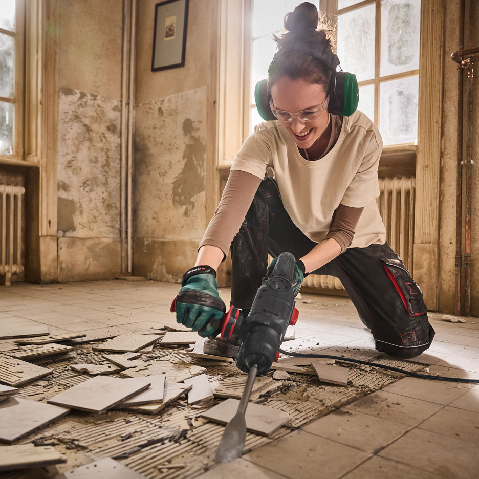 Mulher com protetores auriculares e óculos a usar um martelo perfurador para remover azulejos.