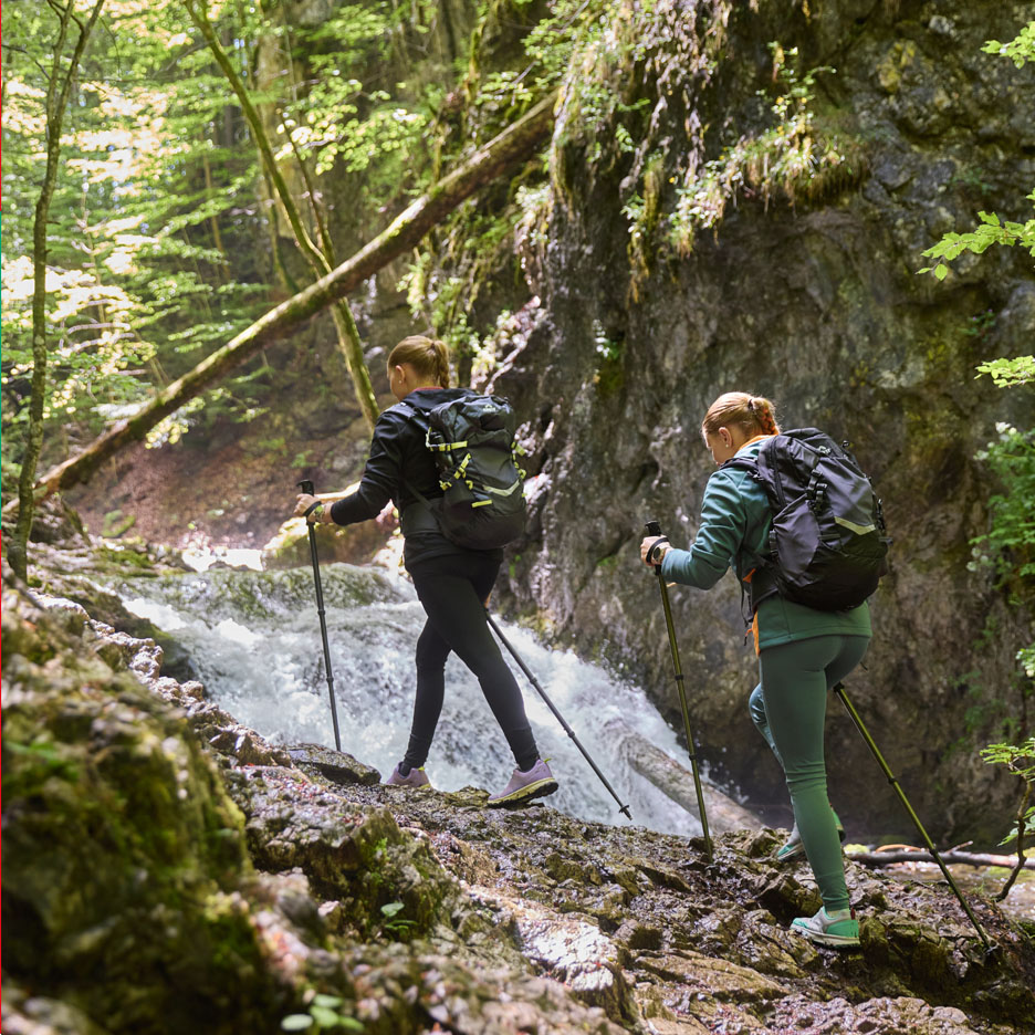 Duas mulheres com mochilas e bastões de caminhada caminham perto de uma cachoeira.