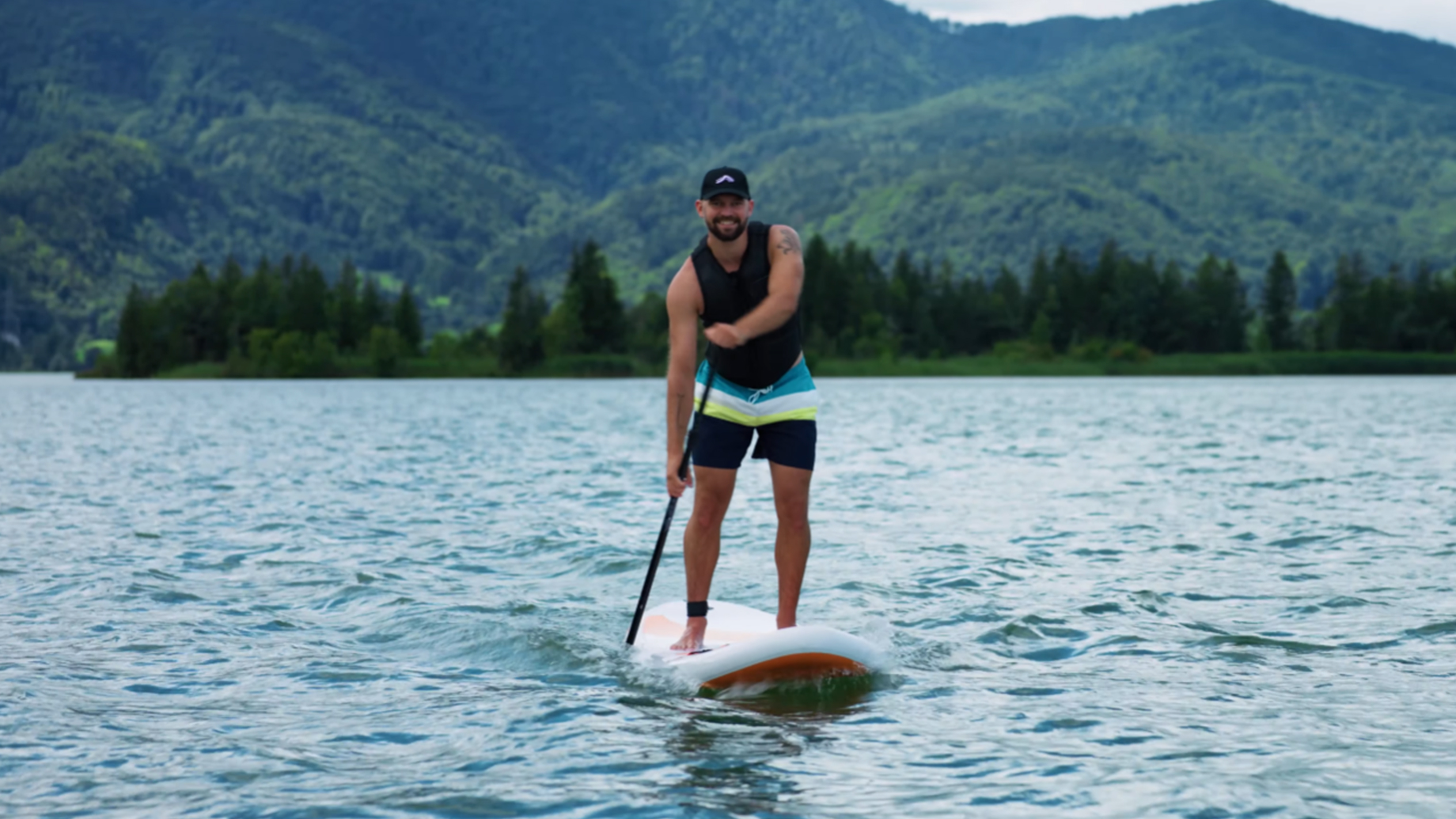 Homem em prancha de paddle surf num lago com montanhas ao fundo.