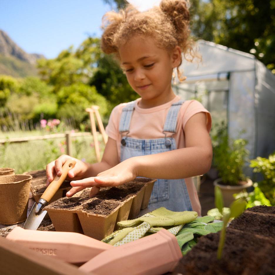Menina a plantar sementes em vasos de turfa com ferramentas de jardinagem e luvas.