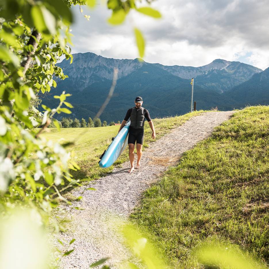 Homem com fato de mergulho e colete salva-vidas a carregar uma prancha de paddleboard numa paisagem montanhosa.