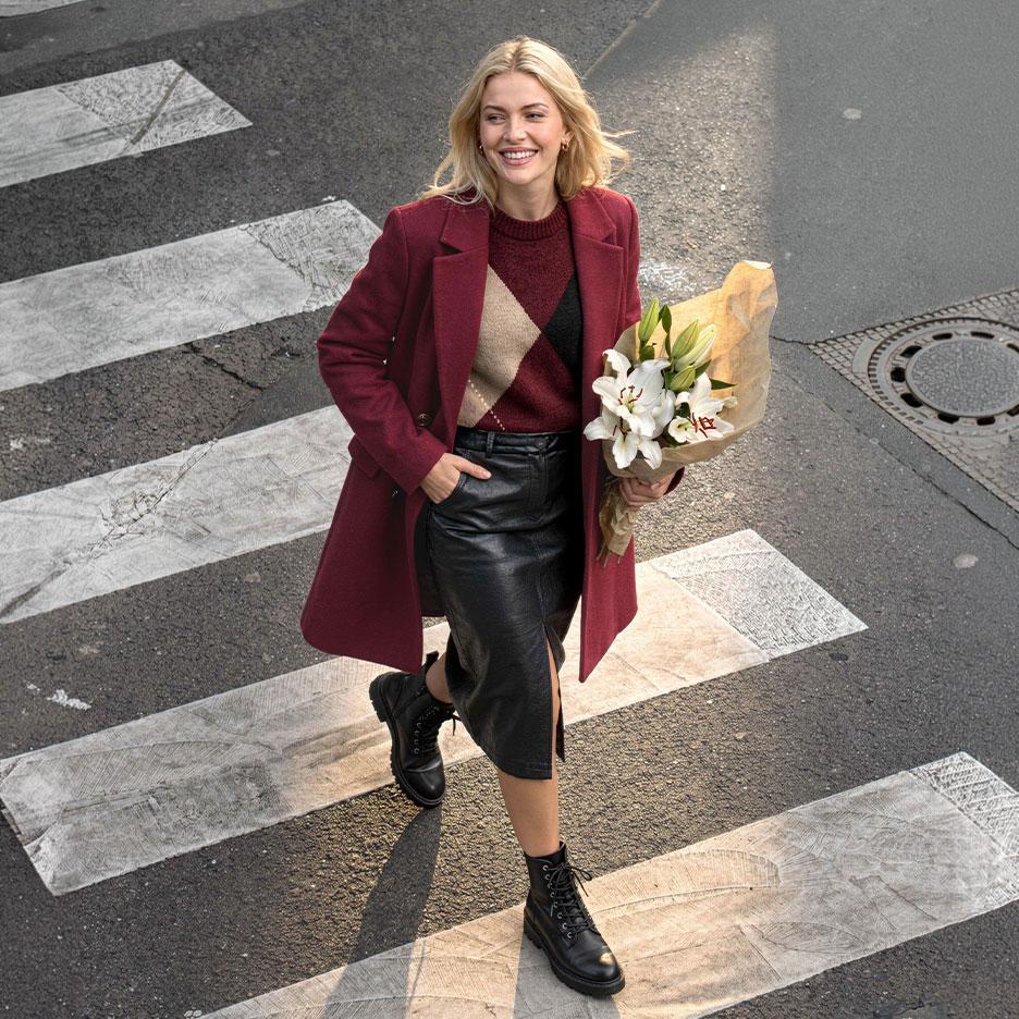 Mulher sorridente com casaco bordô, camisola argyle e saia de couro, a atravessar a rua com um ramo de lírios.