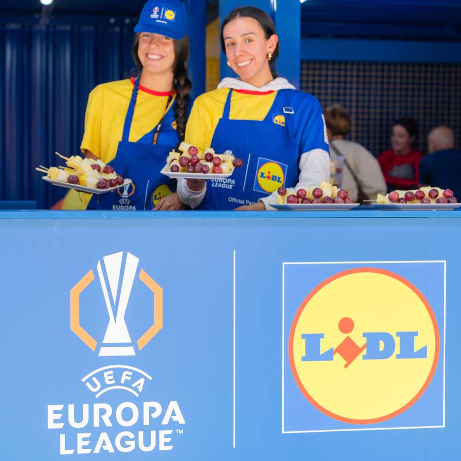Duas mulheres sorridentes com aventais Lidl oferecem espetadas de fruta num evento da UEFA Europa League.