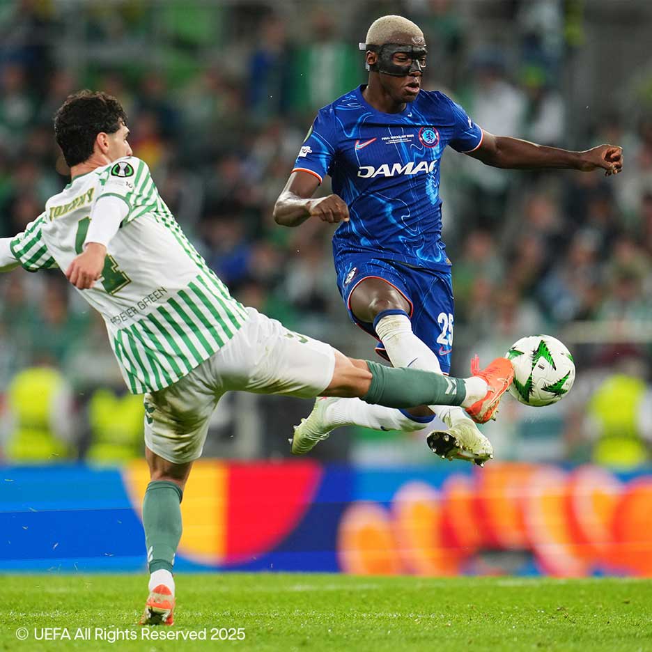 Dois jogadores de futebol lutam pela bola em campo, um de branco e verde, o outro de azul com máscara.