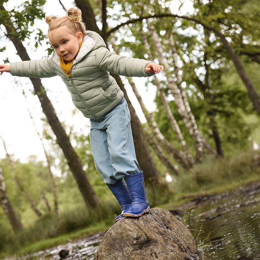 Menina com casaco acolchoado, jeans e galochas a equilibrar-se numa rocha na floresta.