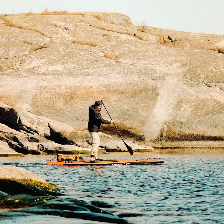 Homem a remar numa prancha de stand-up paddle laranja num lago rochoso.