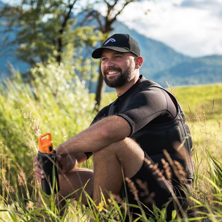 Homem sorridente com boné preto e colete de neoprene, segurando uma garrafa de água laranja.