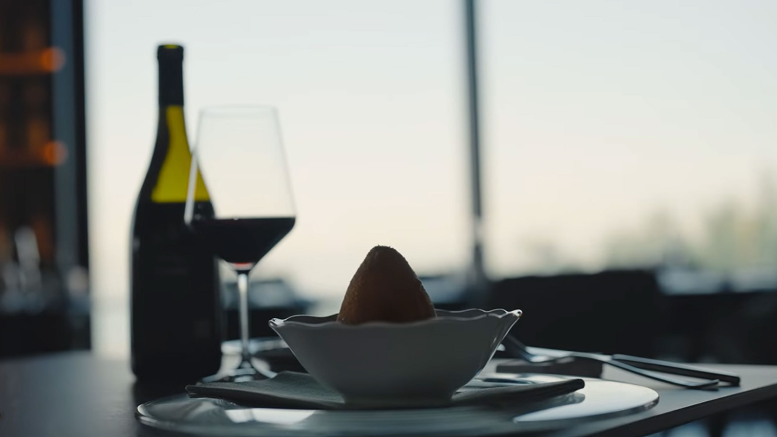 Uma garrafa de vinho tinto e um copo ao lado de um arancino numa tigela sobre uma mesa.