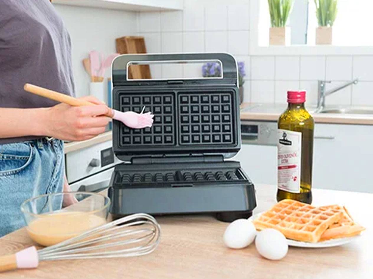 Uma pessoa pincela uma máquina de waffles com um pincel, com ingredientes e waffles prontos na mesa.