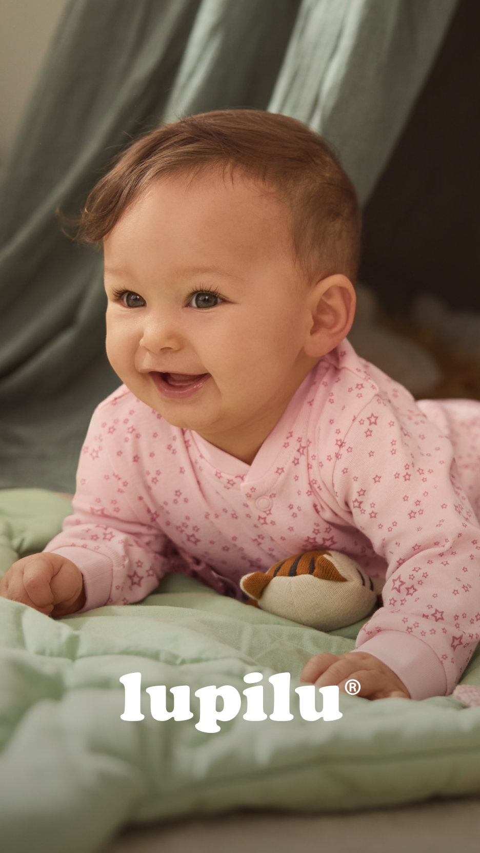 Bebé sorridente deitado de bruços num cobertor verde, vestindo um pijama rosa com estrelas.