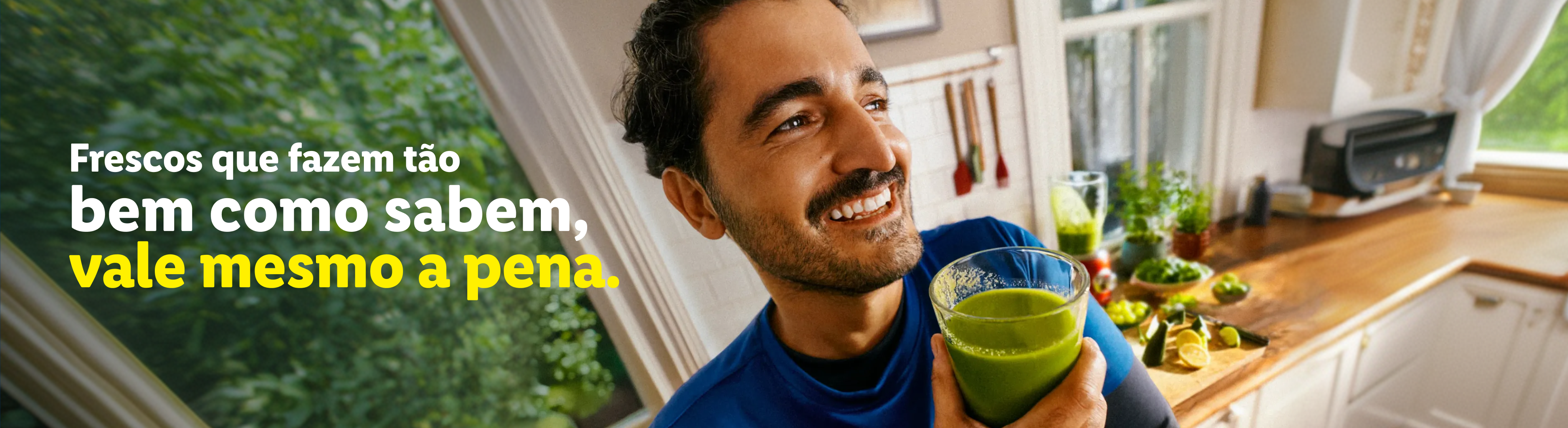 Homem sorridente com um batido verde na cozinha, com o texto: 'Frescos que fazem tão bem como sabem, vale mesmo a pena'.