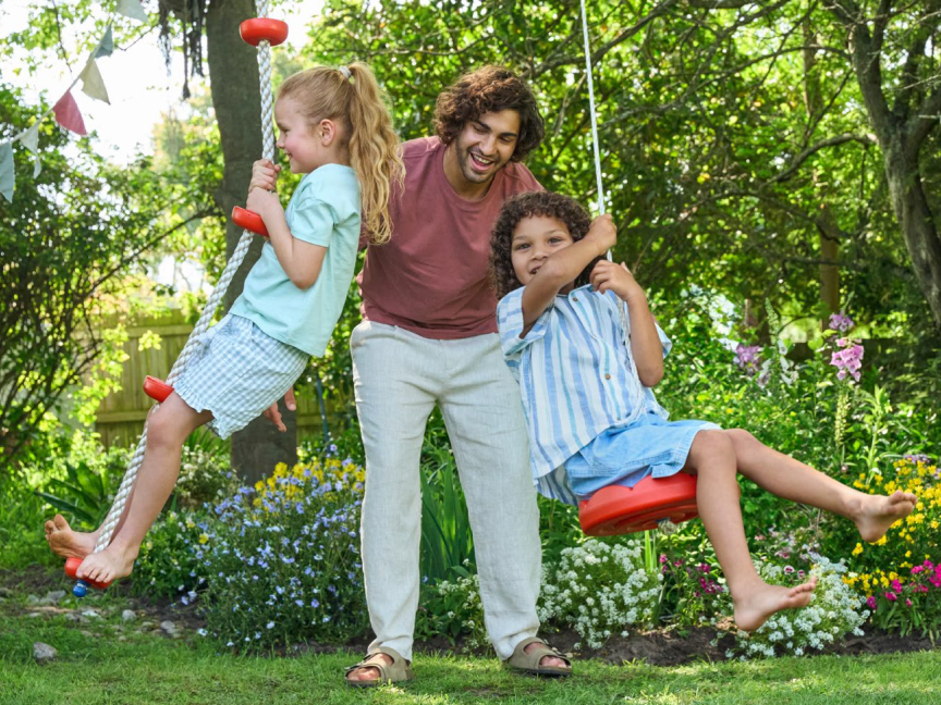 Um pai brinca com seus filhos em um balanço de corda em um jardim florido.