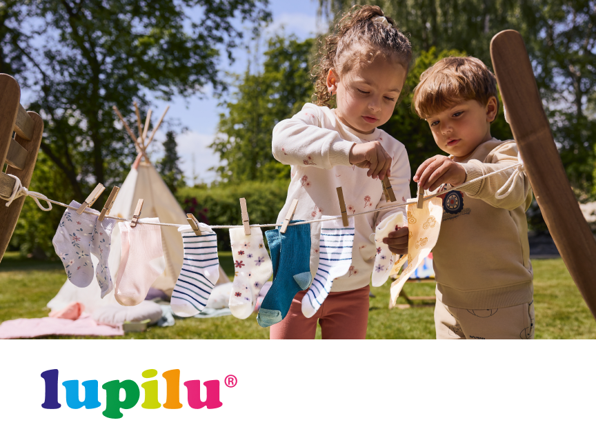 Duas crianças pendurando meias em um varal de brinquedo com uma tenda ao fundo, com o logotipo Lupilu.