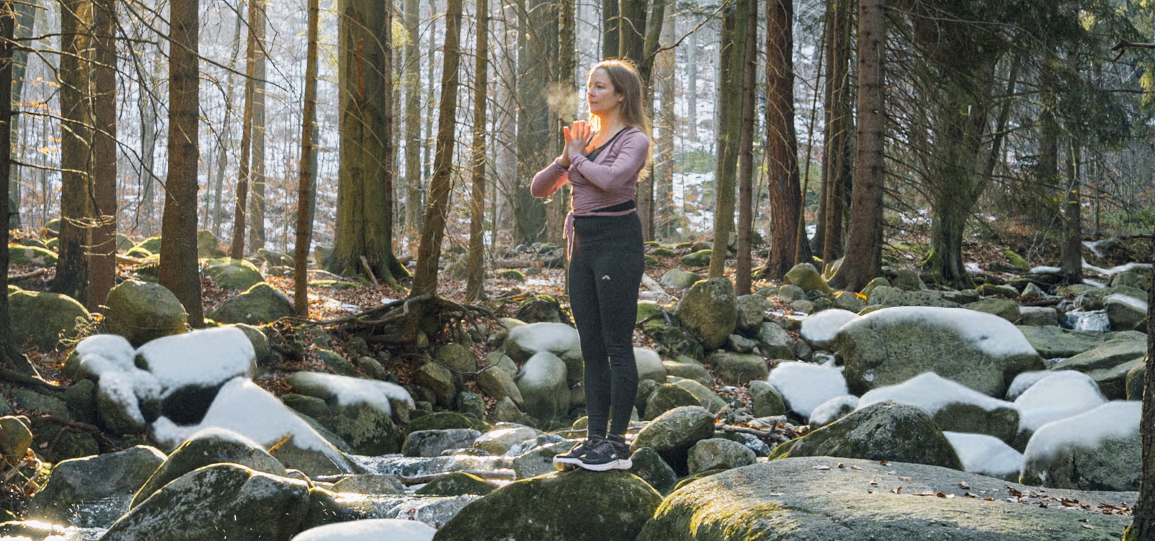 Mulher em roupa desportiva a fazer yoga numa floresta nevada com um riacho.