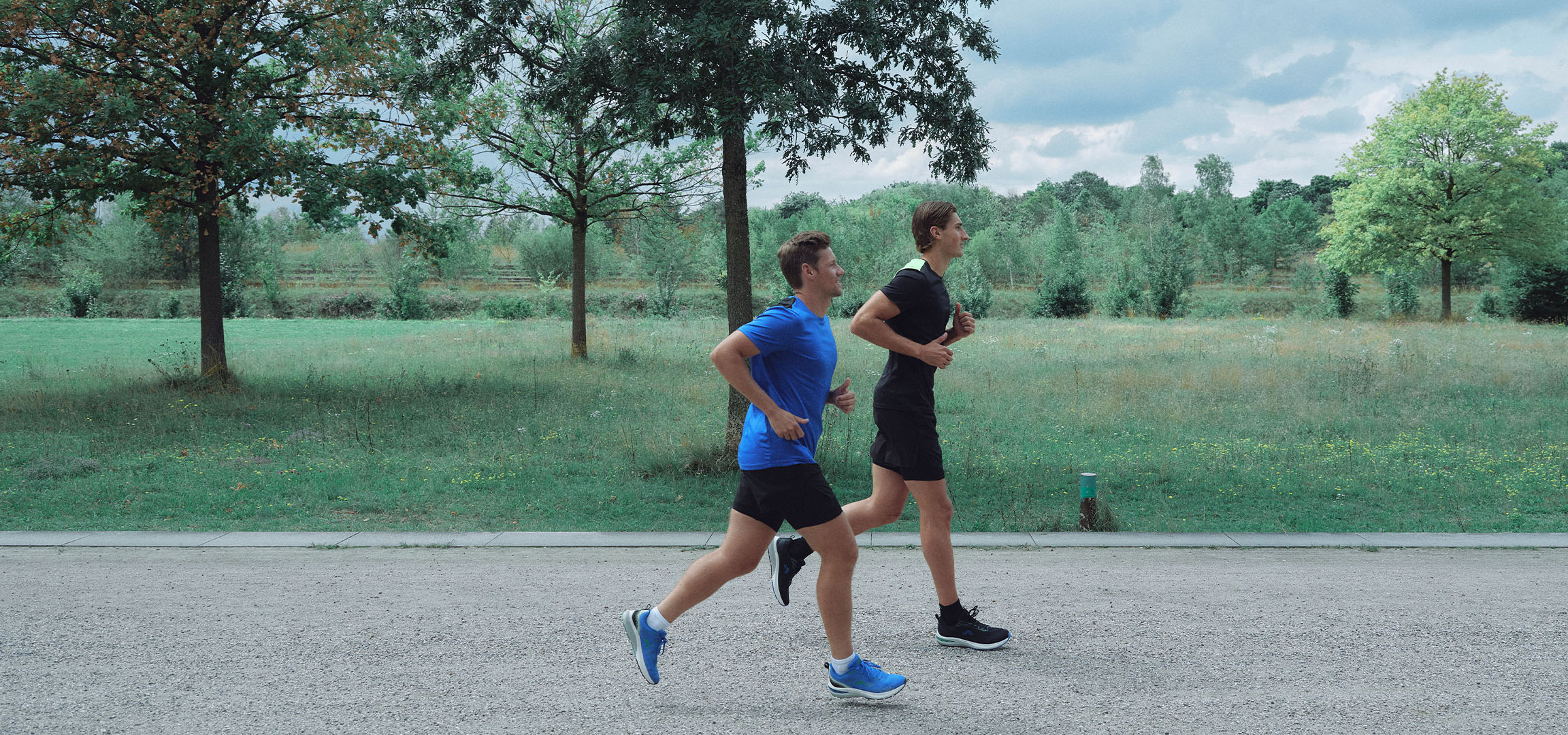 Dois homens a correr num caminho de gravilha num parque, vestidos com roupa desportiva.