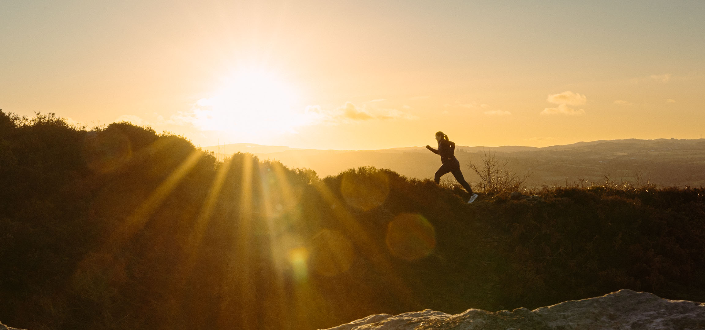 Mulher a correr numa colina ao pôr do sol, com raios de sol e uma paisagem cénica.