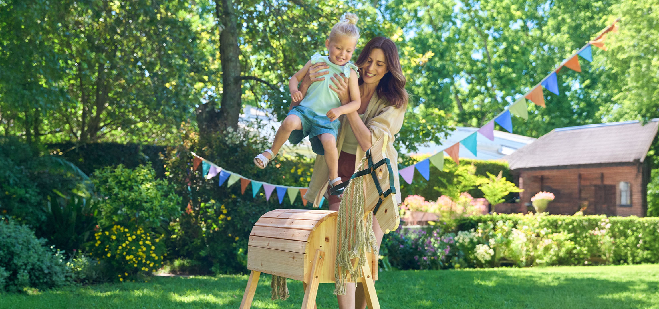 Mãe e criança a brincar com um cavalo de baloiço de madeira num jardim ensolarado.