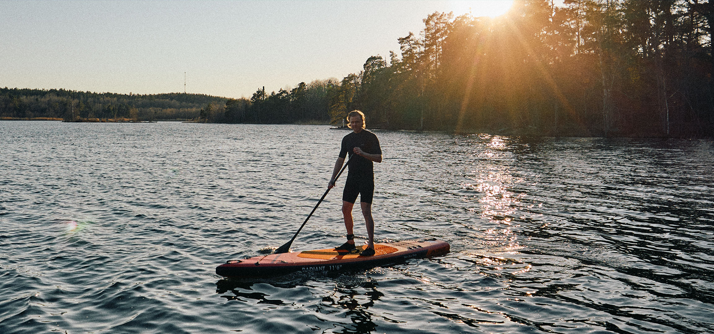 Homem numa prancha de stand up paddle num lago ao pôr do sol.