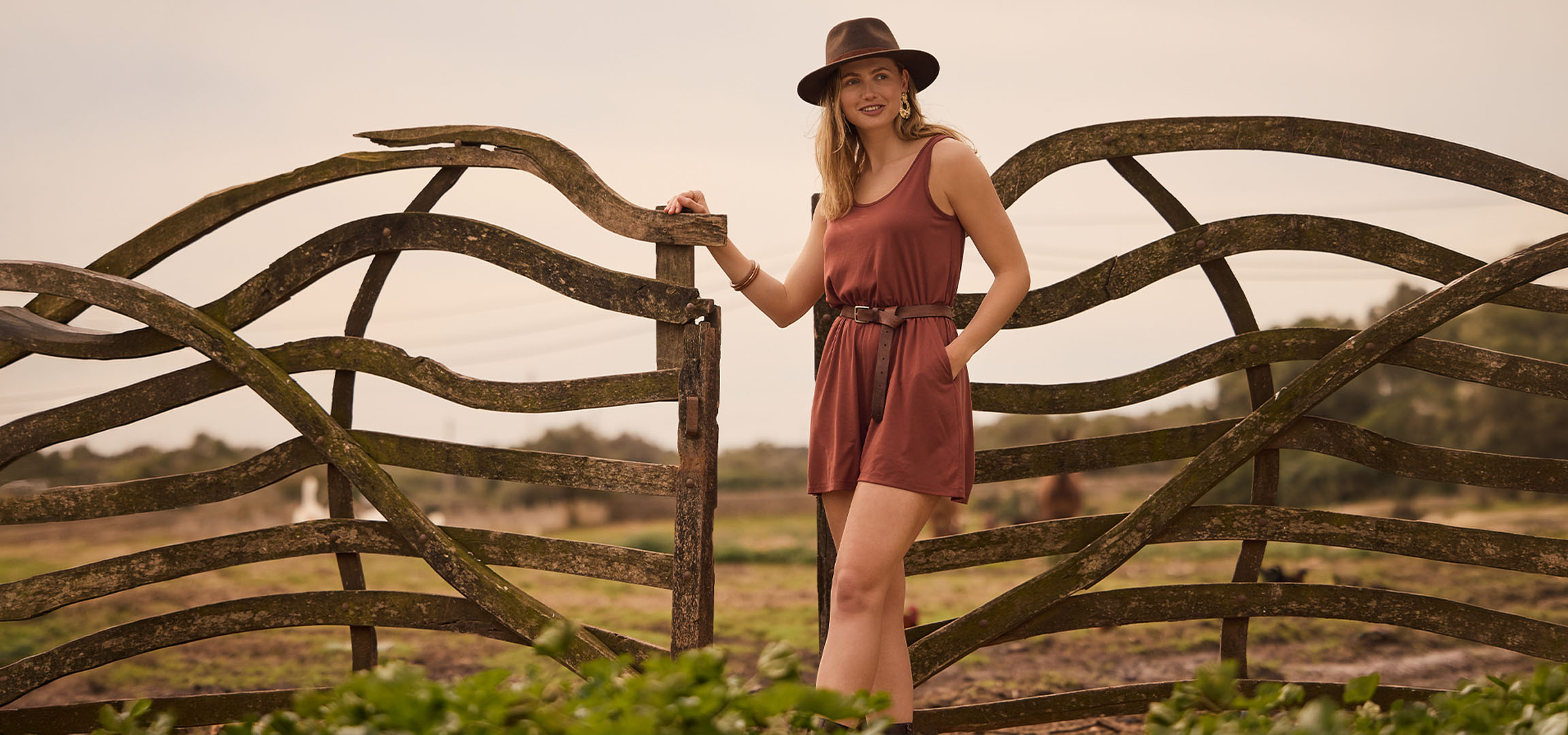 Mulher de vestido e chapéu, encostada a uma cerca de madeira no campo.
