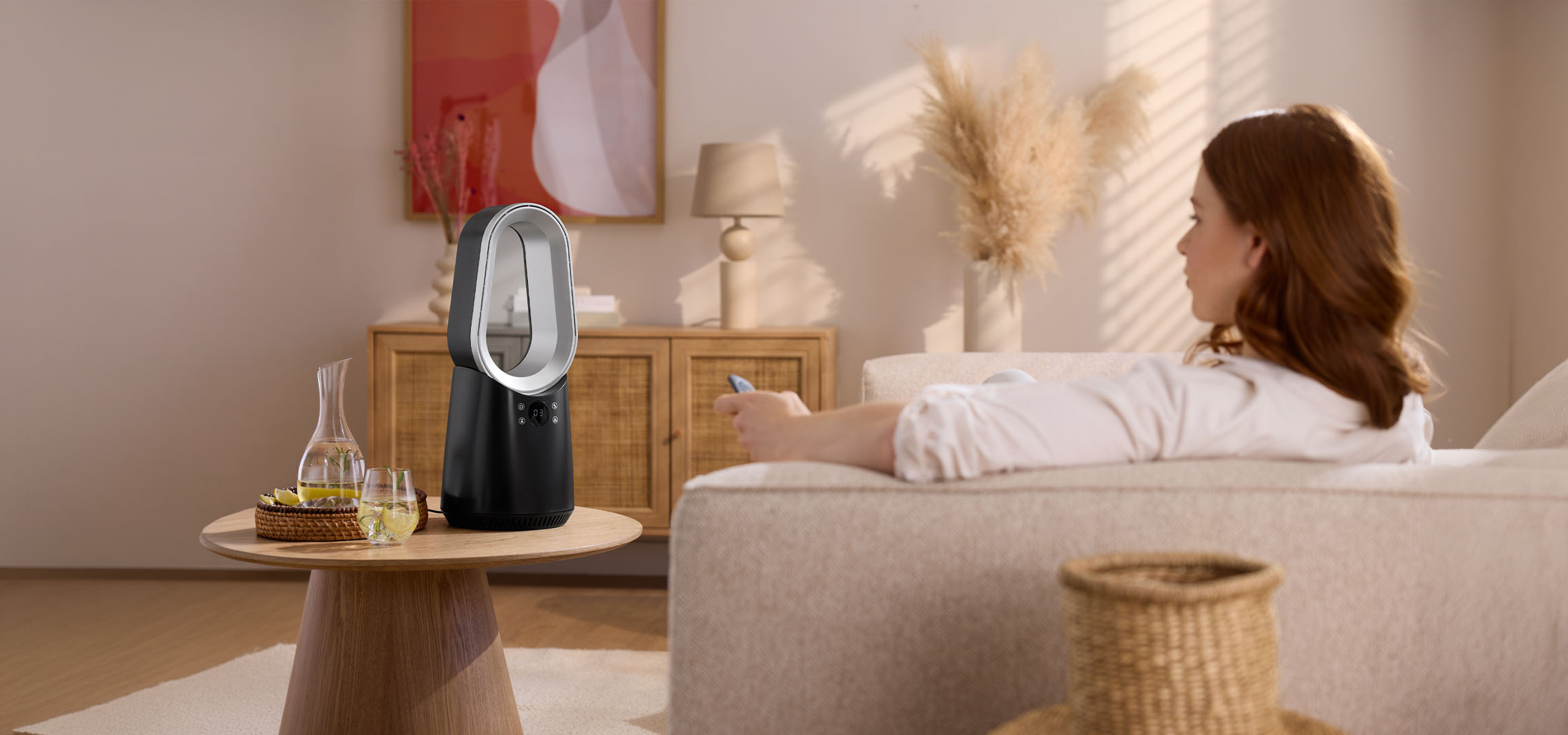 Mulher no sofá com controlo remoto, ventoinha moderna e bebidas na mesa.