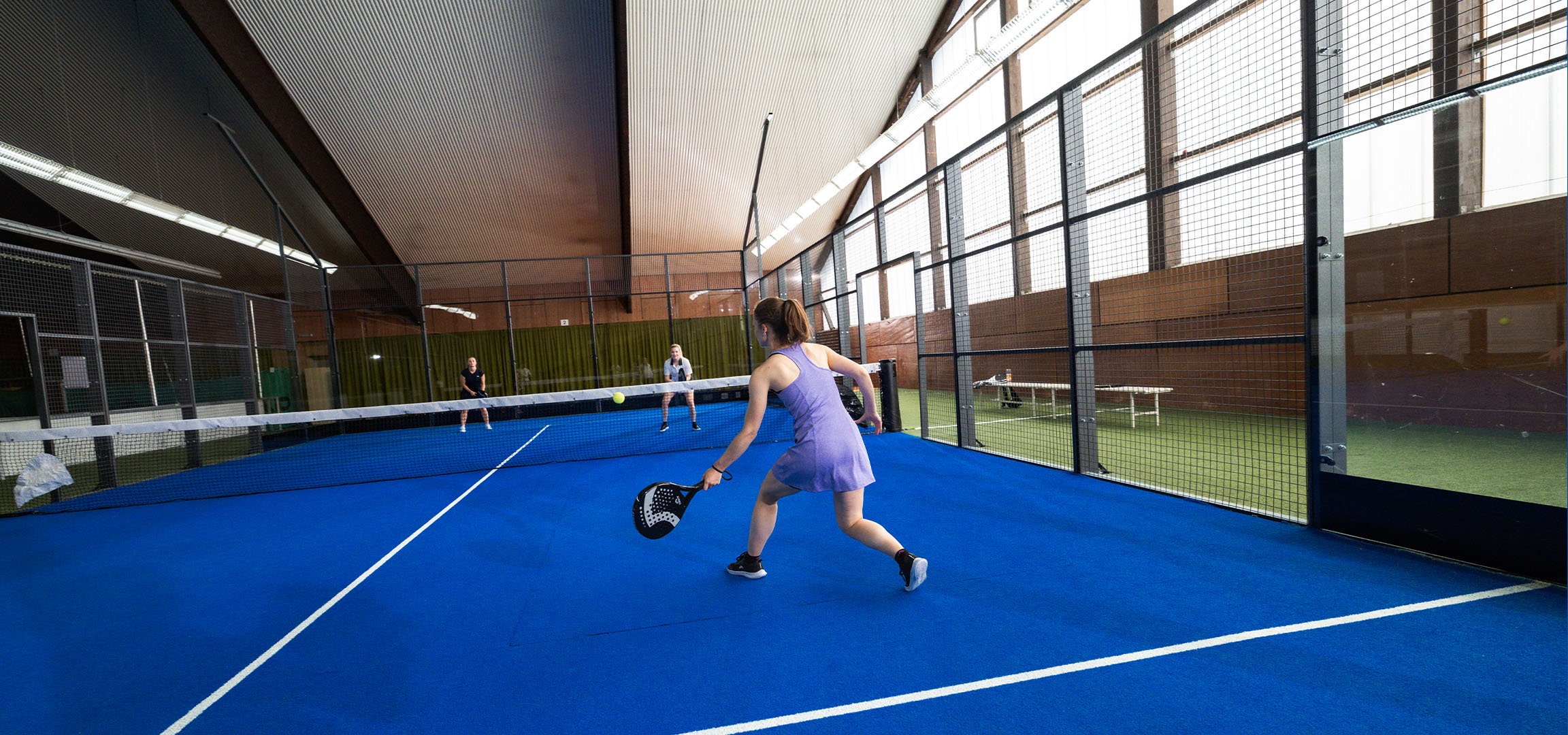Mulher a jogar padel num campo azul, com outros jogadores ao fundo.
