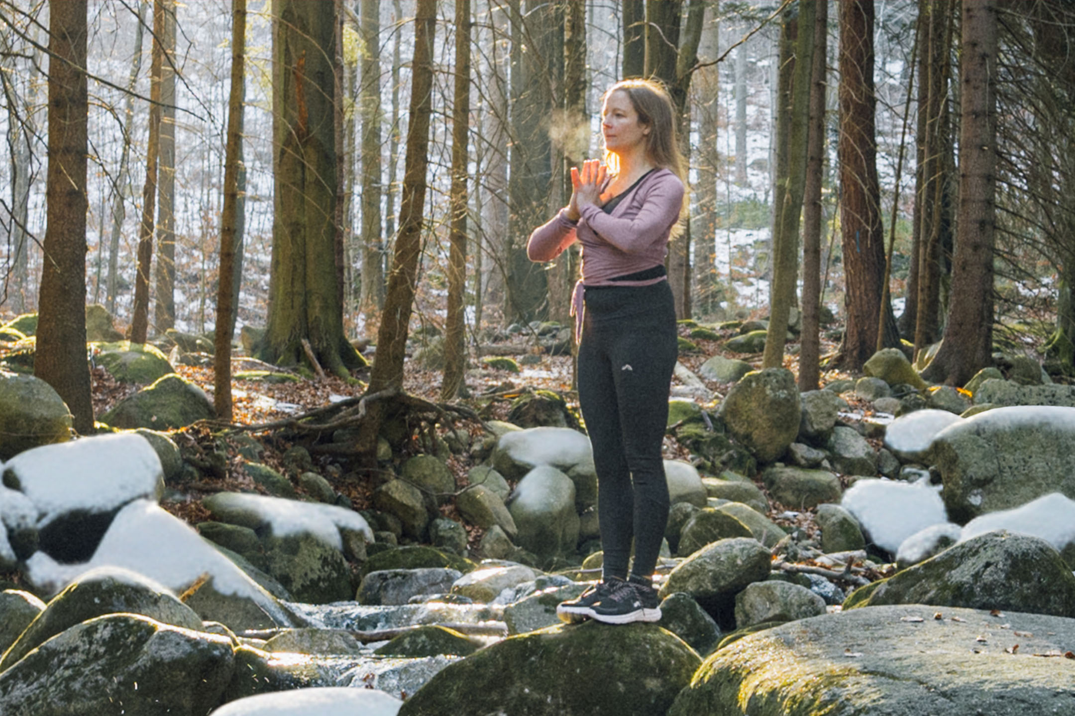 Mulher em roupa desportiva a fazer yoga numa floresta com rochas e árvores nevadas.