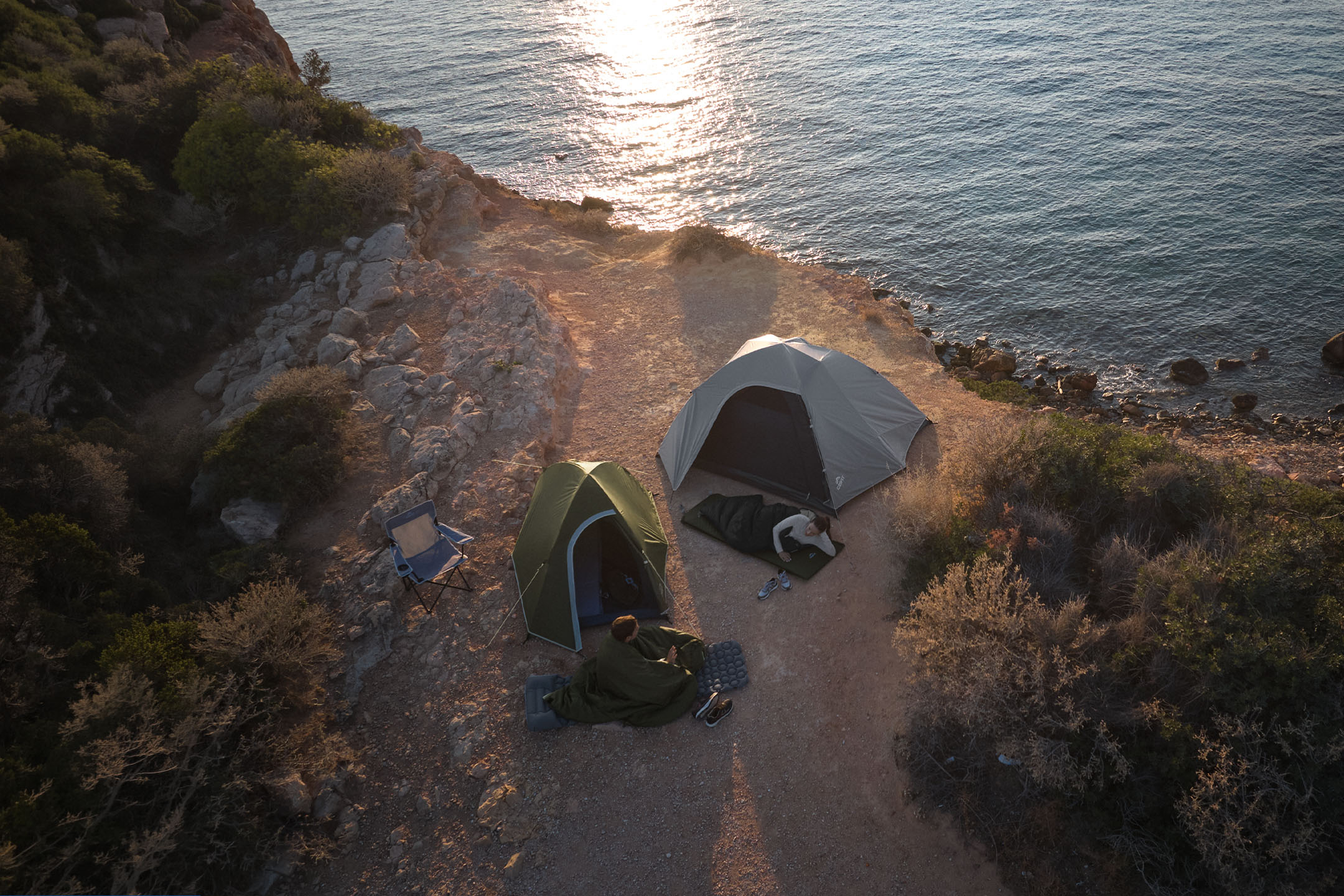 Duas pessoas dormindo em sacos de dormir ao lado de tendas de campismo numa costa rochosa ao pôr do sol.