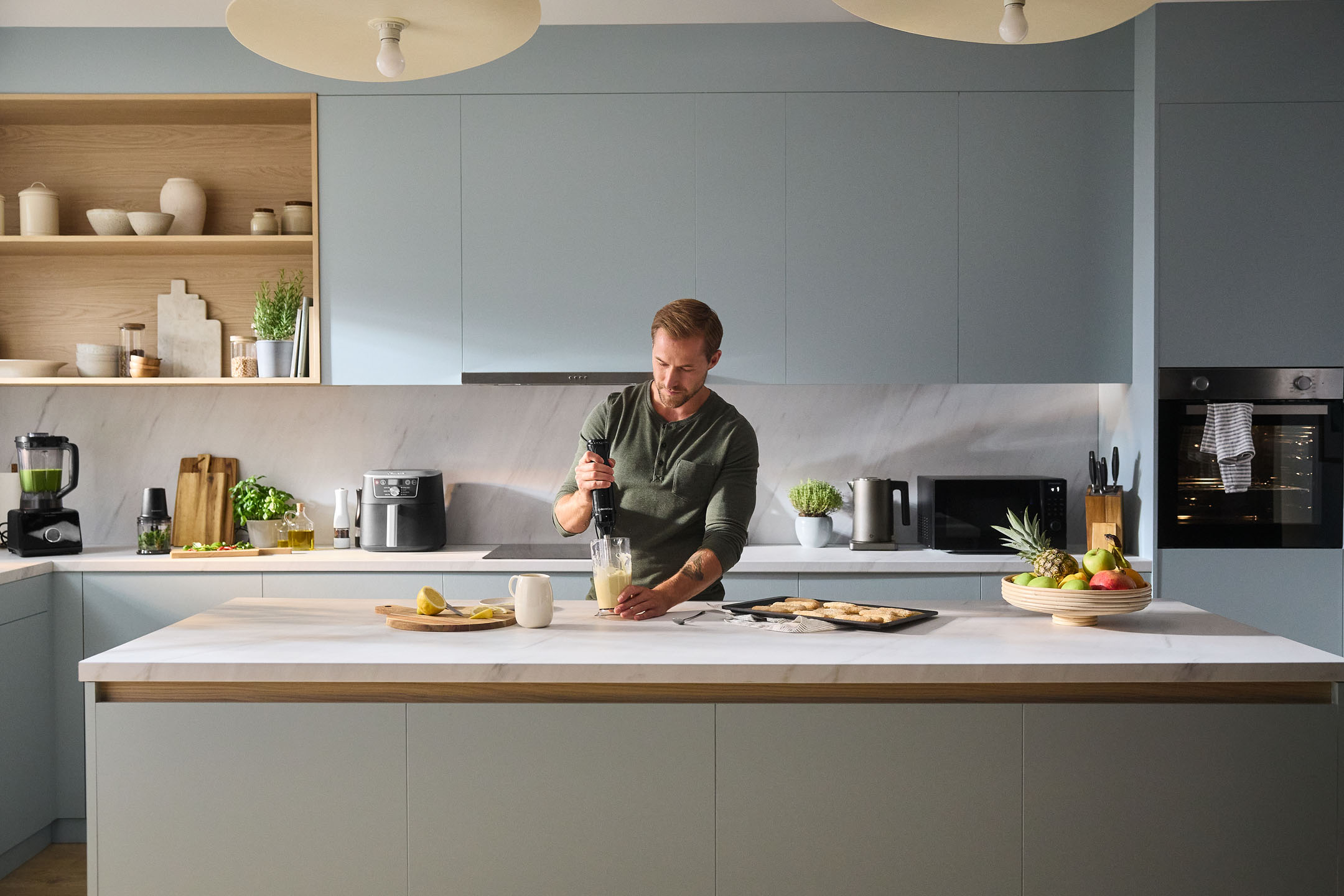 Homem a usar varinha mágica numa cozinha moderna com eletrodomésticos e fruta.