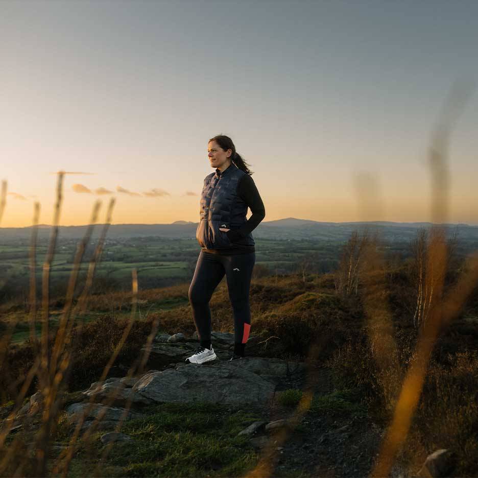 Mulher com colete acolchoado e leggings em pé no topo de uma colina ao pôr do sol.