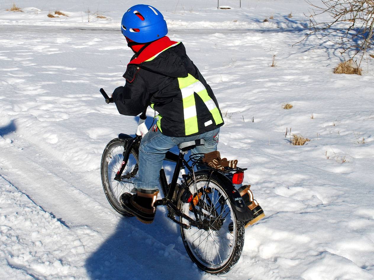 Criança com capacete azul e colete refletor a andar de bicicleta na neve.