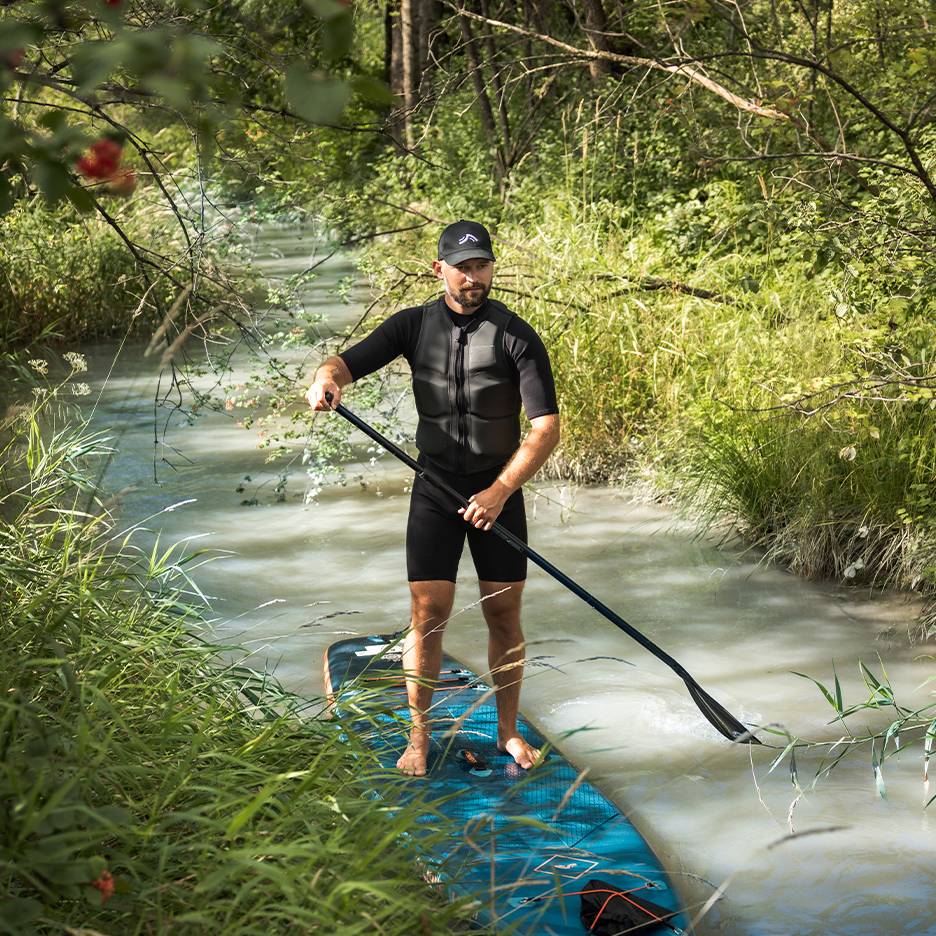 Homem em fato de mergulho e colete de flutuação, em pé numa prancha de paddleboard num riacho.