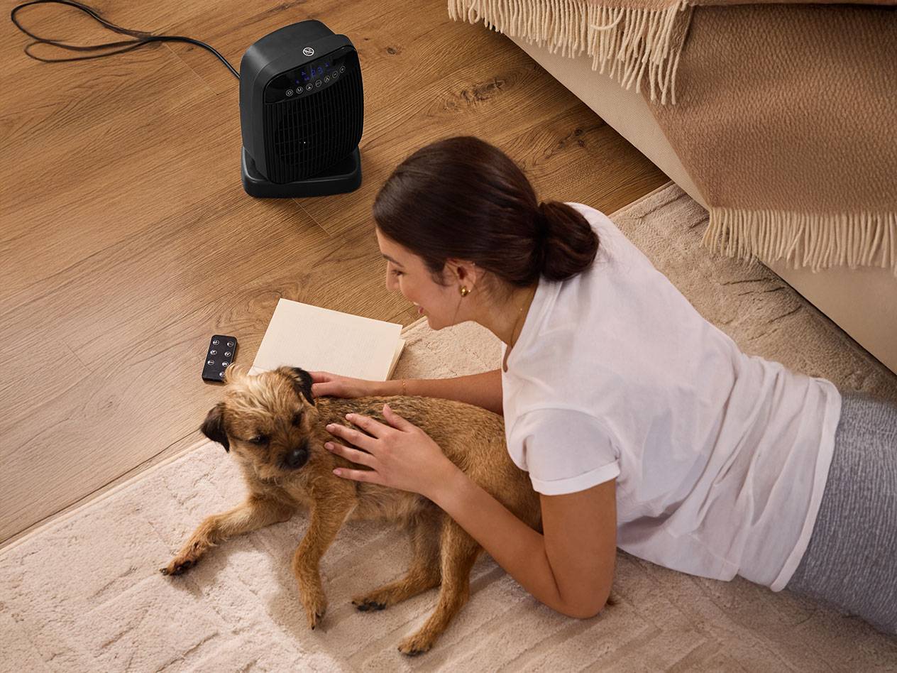 Mulher acariciando um cachorro perto de um aquecedor de ambiente preto no chão.