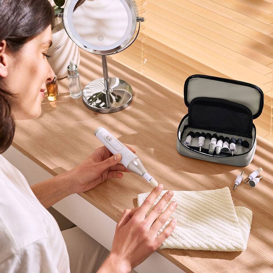 Mulher a usar uma lima elétrica de unhas com acessórios, junto a um espelho e uma bolsa.