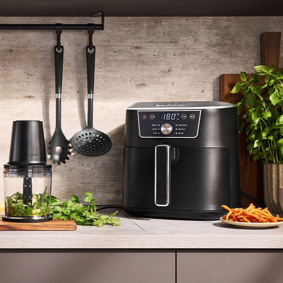 Fritadeira de ar SilverCrest e picador de alimentos numa bancada de cozinha com ervas frescas e batatas fritas de batata doce.