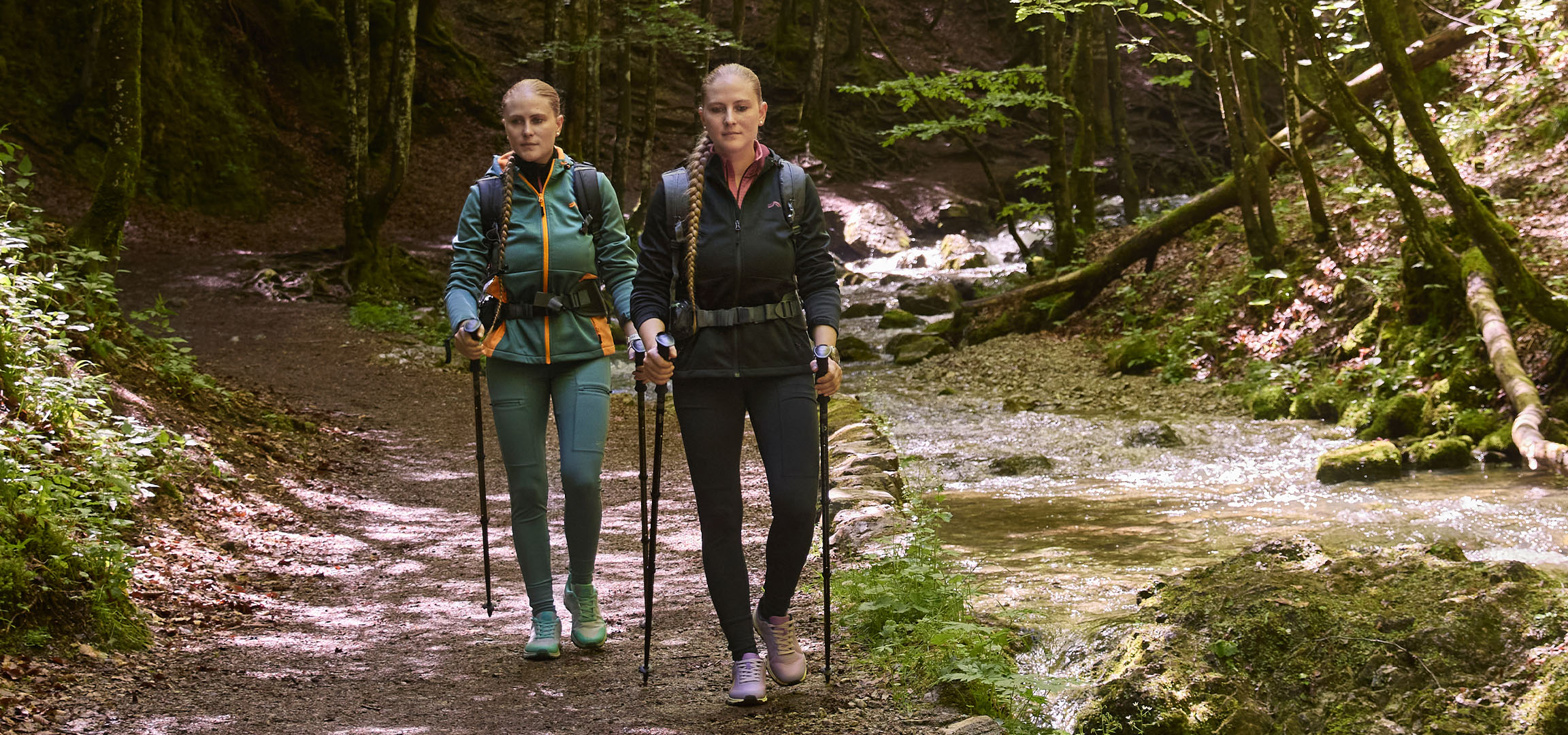 Duas mulheres fazem caminhada ao longo de um riacho numa floresta, usando bastões de caminhada.
