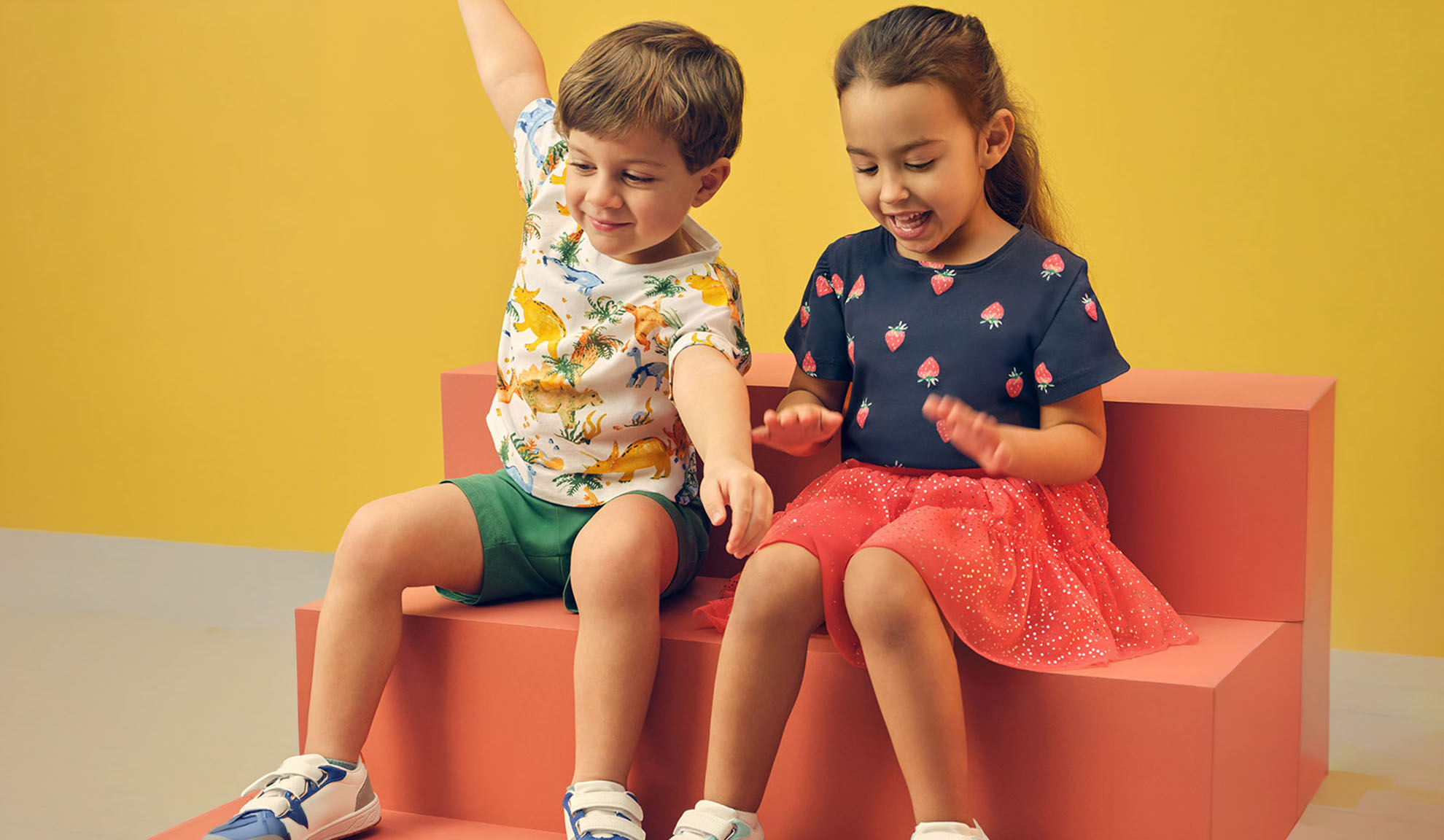 Duas crianças com roupas de verão, um menino com t-shirt de dinossauro e uma menina com t-shirt de morango.