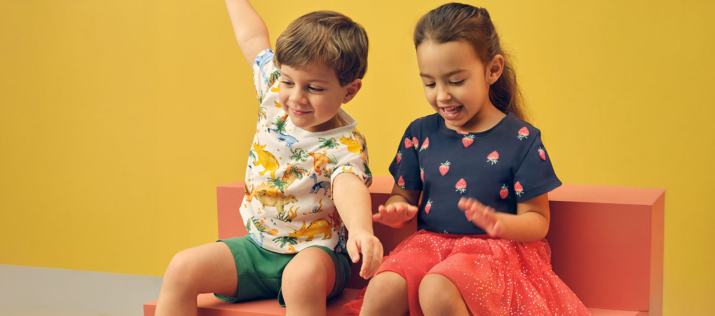 Duas crianças, um menino e uma menina, sentadas num banco rosa com roupas de verão.