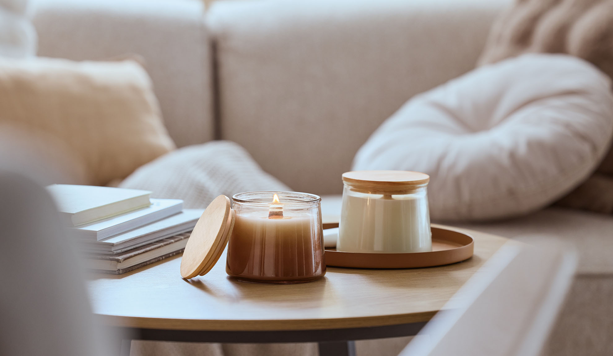 Duas velas perfumadas numa mesa de madeira, uma acesa, com um sofá acolhedor ao fundo.