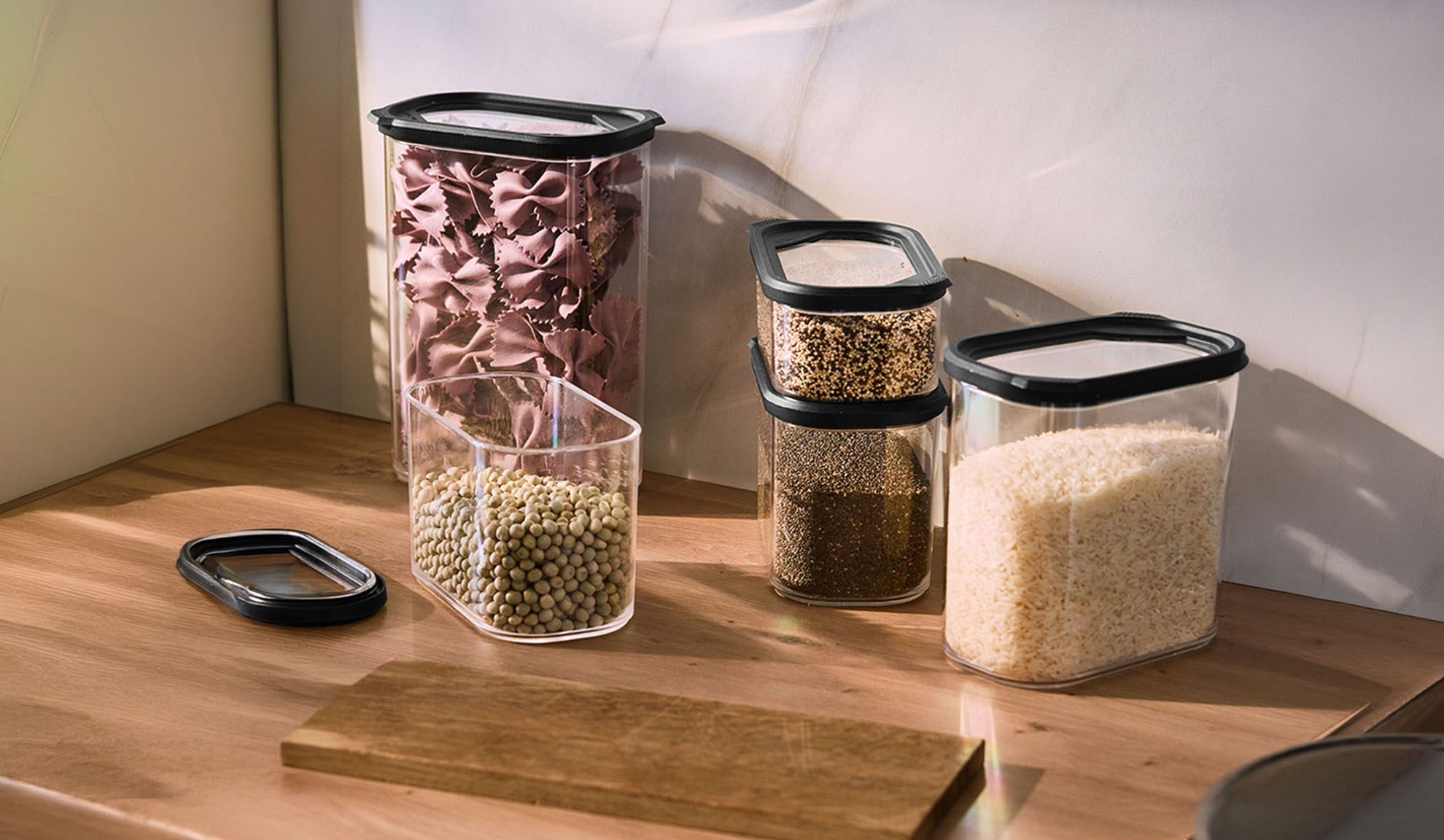 Recipientes transparentes de armazenamento de cozinha com massa, lentilhas, sementes e arroz numa bancada de madeira.